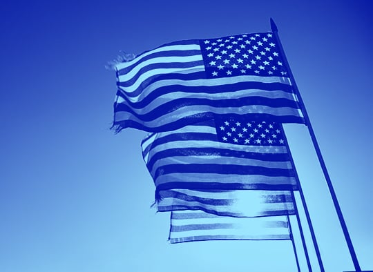 four-american-flags-fly-in-breeze-with-sun-behind-blue-duotone